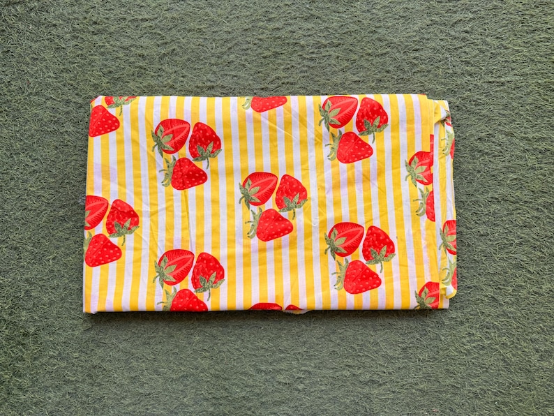 May include: Folded fabric featuring a repeating pattern of red strawberries with green stems on a yellow and white striped background. The fabric is folded neatly, showcasing the design.