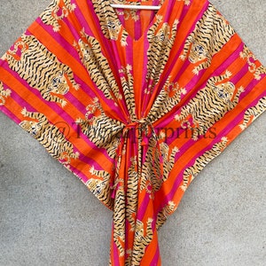May include: A vibrant orange and pink striped kaftan with a V-neck and a tie closure. The fabric is adorned with a repeating pattern of tiger illustrations in black and tan. The kaftan is hanging on a white hanger.