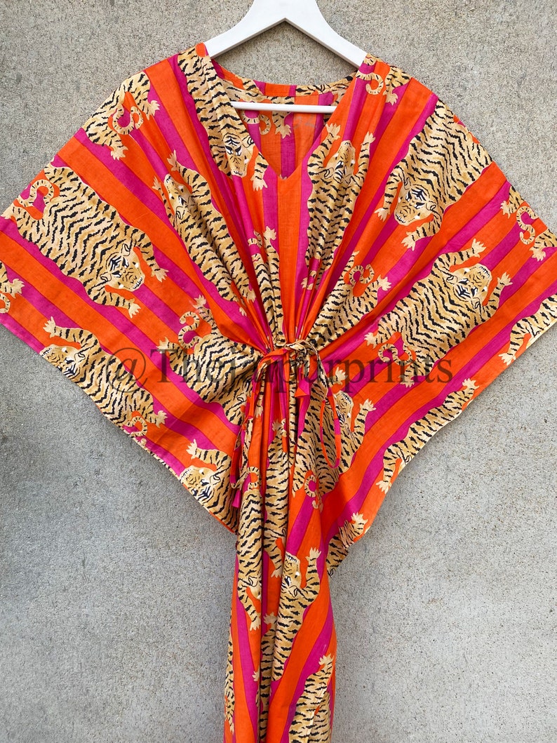 May include: A vibrant orange and pink striped kaftan with a V-neck and a tie closure. The fabric is adorned with a repeating pattern of tiger illustrations in black and tan. The kaftan is hanging on a white hanger.
