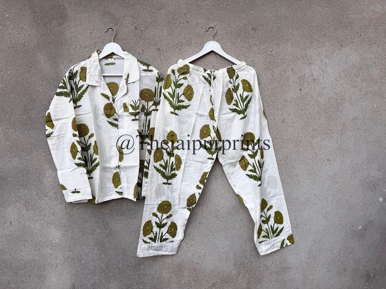 May include: White pajama set with a green floral print. The set includes a button-down shirt and matching pants, both hanging on a white hanger. The floral design is repeated throughout the shirt and pants.