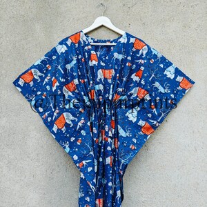 May include: A blue cotton kaftan with a white and orange print of various animals, including polar bears, reindeer, and foxes. The kaftan is hanging on a white hanger against a gray wall.