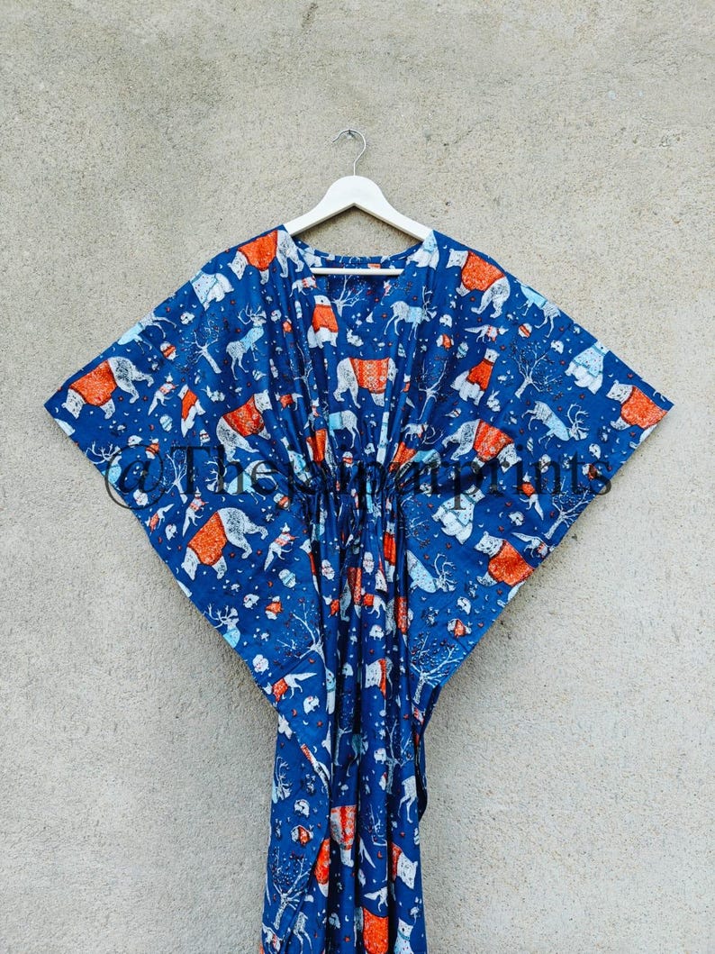 May include: A blue cotton kaftan with a white and orange print of various animals, including polar bears, reindeer, and foxes. The kaftan is hanging on a white hanger against a gray wall.