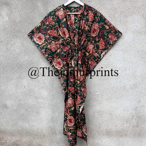 May include: A green cotton kaftan with a floral print in shades of red, orange, and yellow. The kaftan has a V-neckline and a tie at the waist. The kaftan is hanging on a white hanger against a gray wall. @The_Print_Prints