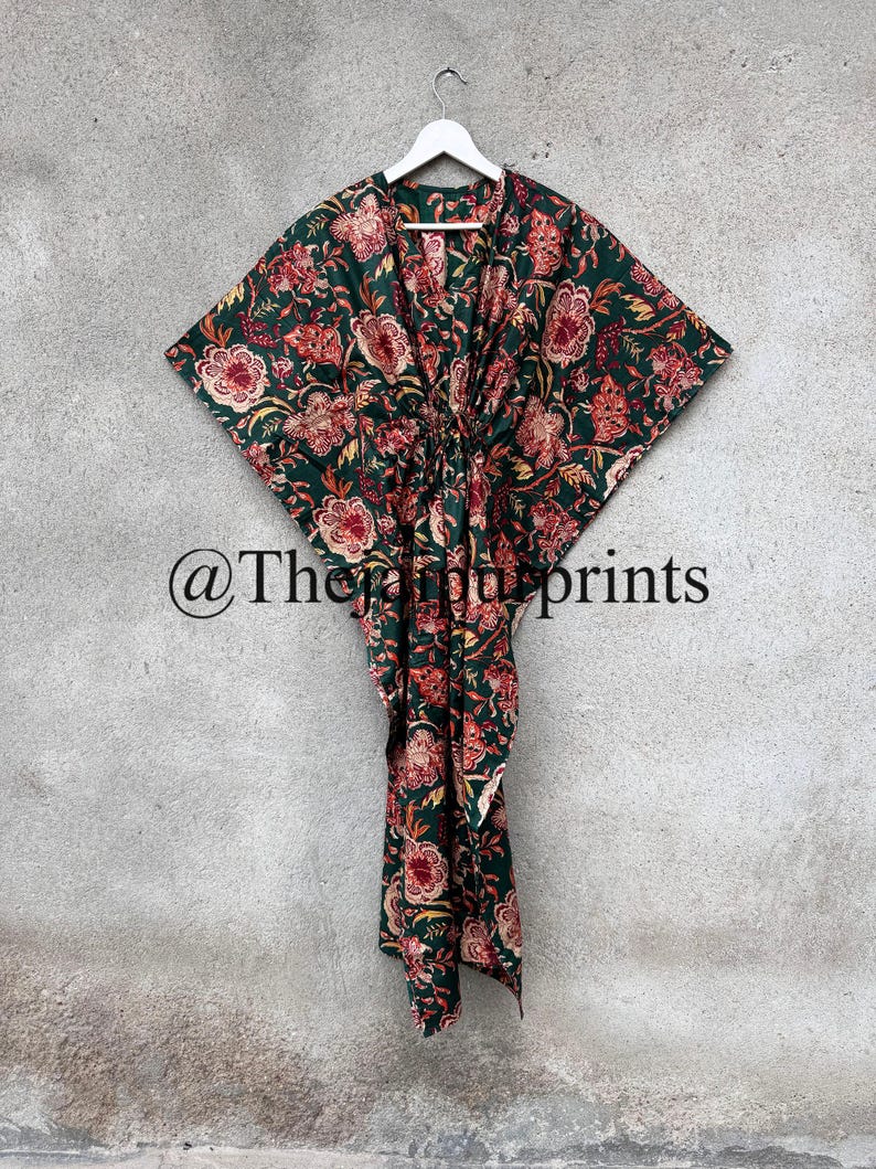 May include: A green cotton kaftan with a floral print in shades of red, orange, and yellow. The kaftan has a V-neckline and a tie at the waist. The kaftan is hanging on a white hanger against a gray wall. @The_Print_Prints