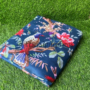 Op de afbeelding: Een donkerblauwe stof met een bloemenprint met rode, roze en witte bloemen en een kleurrijke vogel. De stof is gevouwen en ligt op een groene ondergrond.