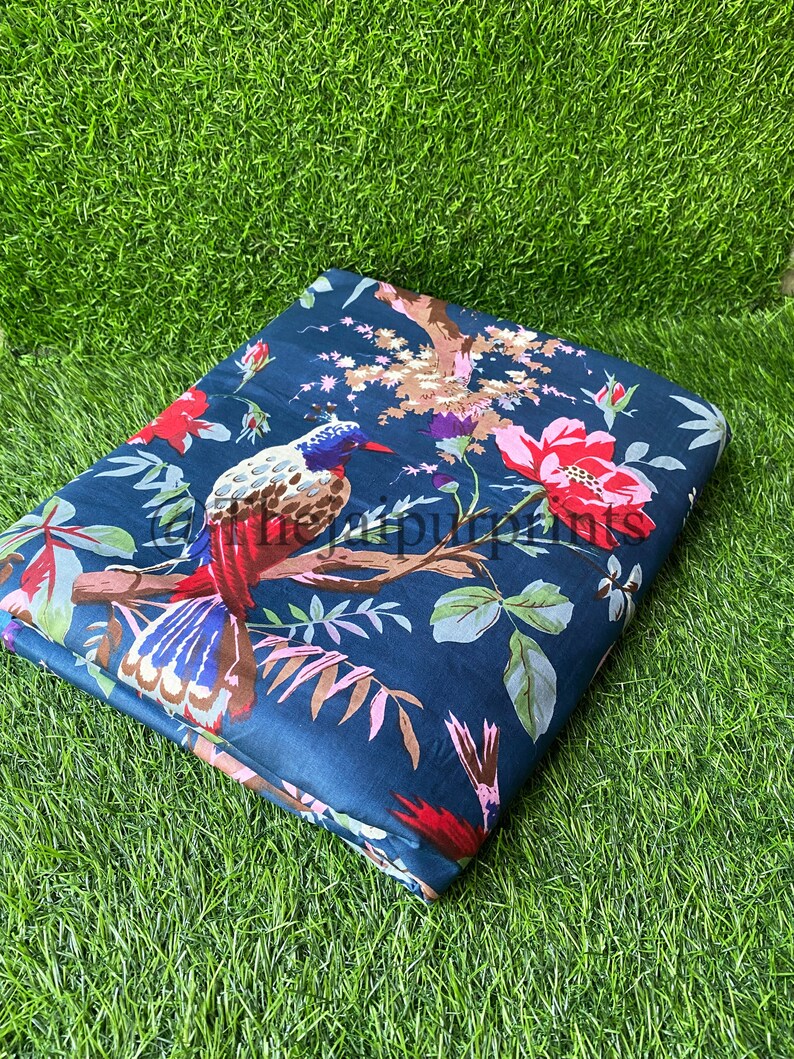 Op de afbeelding: Een donkerblauwe stof met een bloemenprint met rode, roze en witte bloemen en een kleurrijke vogel. De stof is gevouwen en ligt op een groene ondergrond.