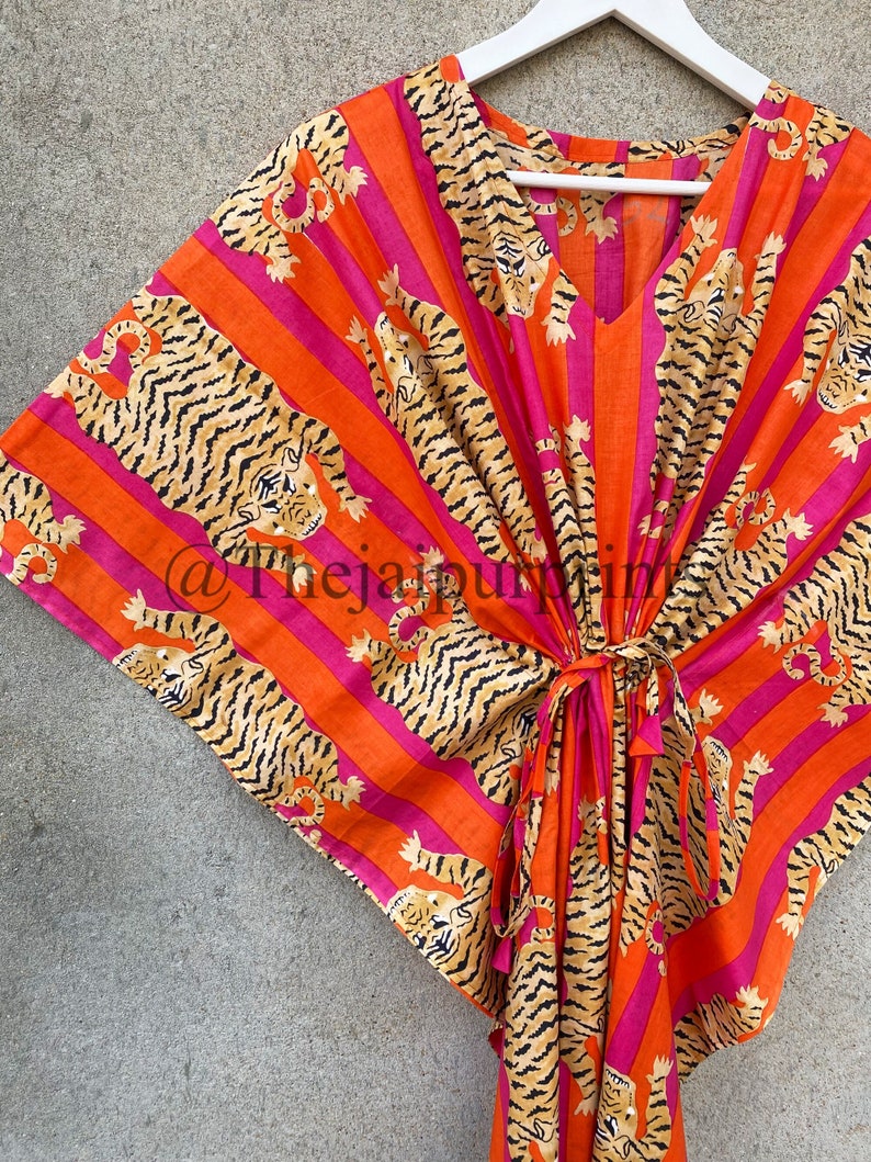 May include: A vibrant orange and pink kaftan with a tiger print. The kaftan features vertical stripes and a repeating pattern of tigers in a relaxed pose. It has a V-neck and a tie at the waist. The garment is hanging on a white hanger.