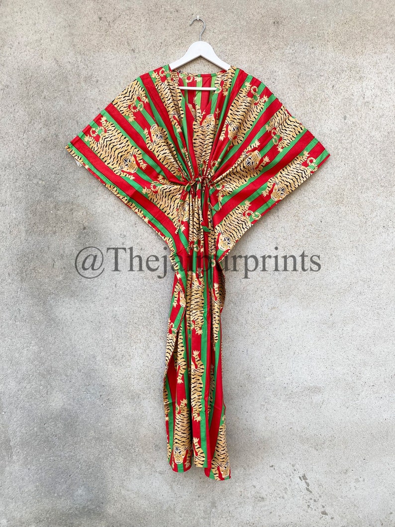 May include: A red, green, and gold patterned kaftan dress with a tiger stripe design. The dress is hanging on a white hanger against a gray background. The text "@Thejapurprints" is visible.
