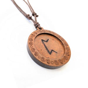 Puede incluir: Un collar con un colgante de madera y un cordón de cuero marrón. El colgante es redondo y presenta un símbolo de runa tallado con un borde decorativo.