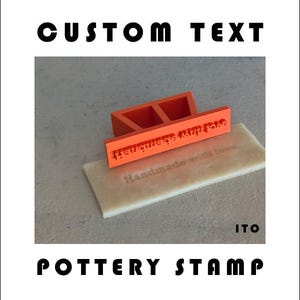 Peut inclure: Un tampon de poterie orange avec le texte "Handmade with love" et "Heritage Mugs & More" sur une base rectangulaire. Les mots "Custom Text" et "Pottery Stamp" sont imprimés au-dessus et en dessous du tampon.