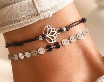 May include: A set of two anklets worn together. One is a black cord featuring a silver-toned lotus flower charm. The second is a delicate silver-toned chain composed of small, flat, circular metallic discs linked together in a row.