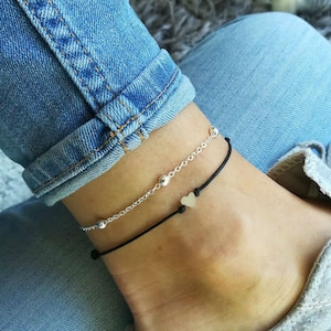 Può includere: Due cavigliere su una caviglia. Una cavigliera è una catena d'argento con piccole perline d'argento. L'altra cavigliera è un cordoncino nero con un piccolo ciondolo a forma di cuore d'argento.