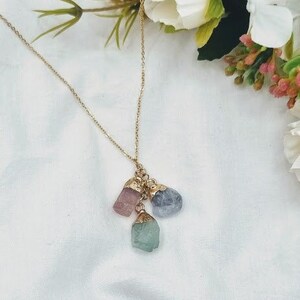 May include: A gold chain necklace with a pendant featuring three raw crystal stones: a light pink stone, a light blue stone, and a light green stone.