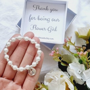 May include: A white pearl bracelet with a silver charm that has the letter "J" on it. The bracelet is in a small white box with the text "Thank you for being our Flower Girl" and the website "artBeadsEtsy.com".