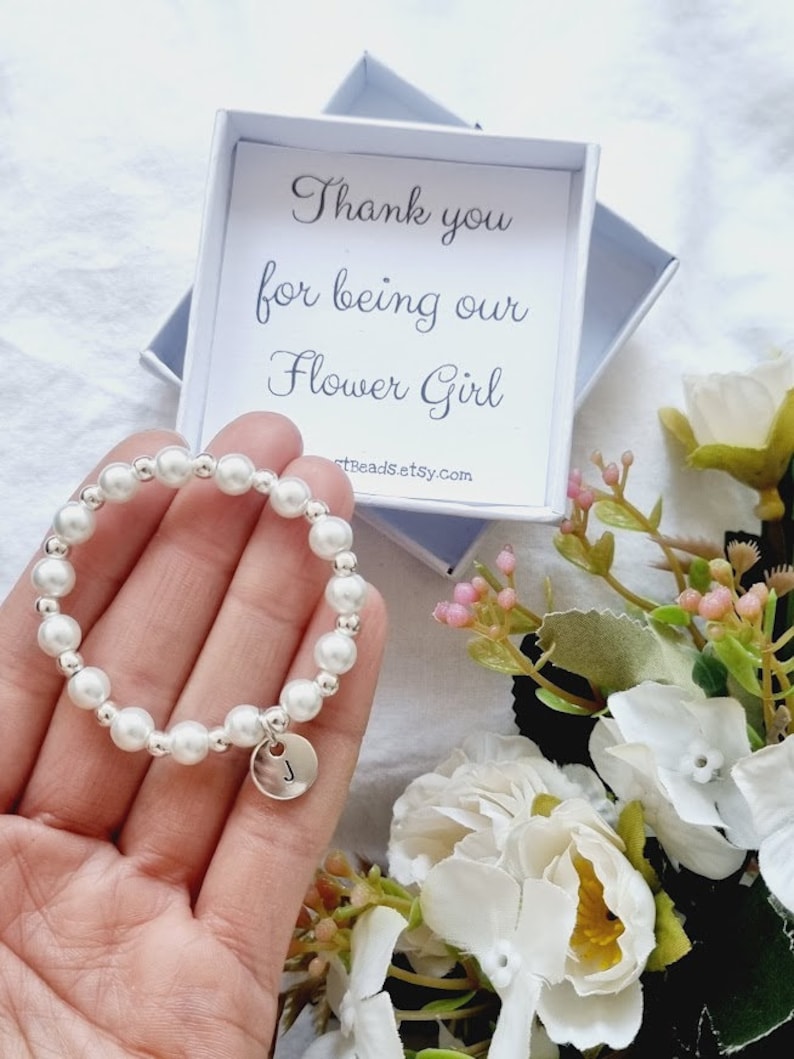 May include: A white pearl bracelet with a silver charm that has the letter "J" on it. The bracelet is in a small white box with the text "Thank you for being our Flower Girl" and the website "artBeadsEtsy.com".