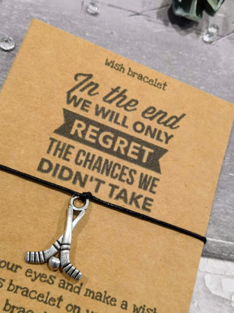 May include: A black string bracelet with a silver hockey stick charm. The bracelet card reads "In the end we will only regret the chances we didn't take".