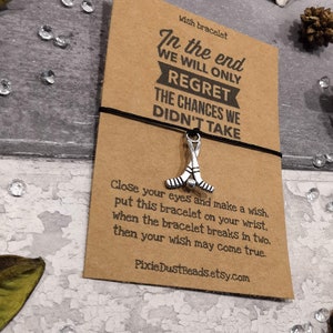 May include: A brown card with the text "In the end we will only regret the chances we didn't take" and a silver hockey stick charm attached to a black string. The card also includes the text "Wish bracelet" and "Close your eyes and make a wish. Put this bracelet on your wrist. When the bracelet breaks in two, then your wish may come true. PixieDustBeads.etsy.com."
