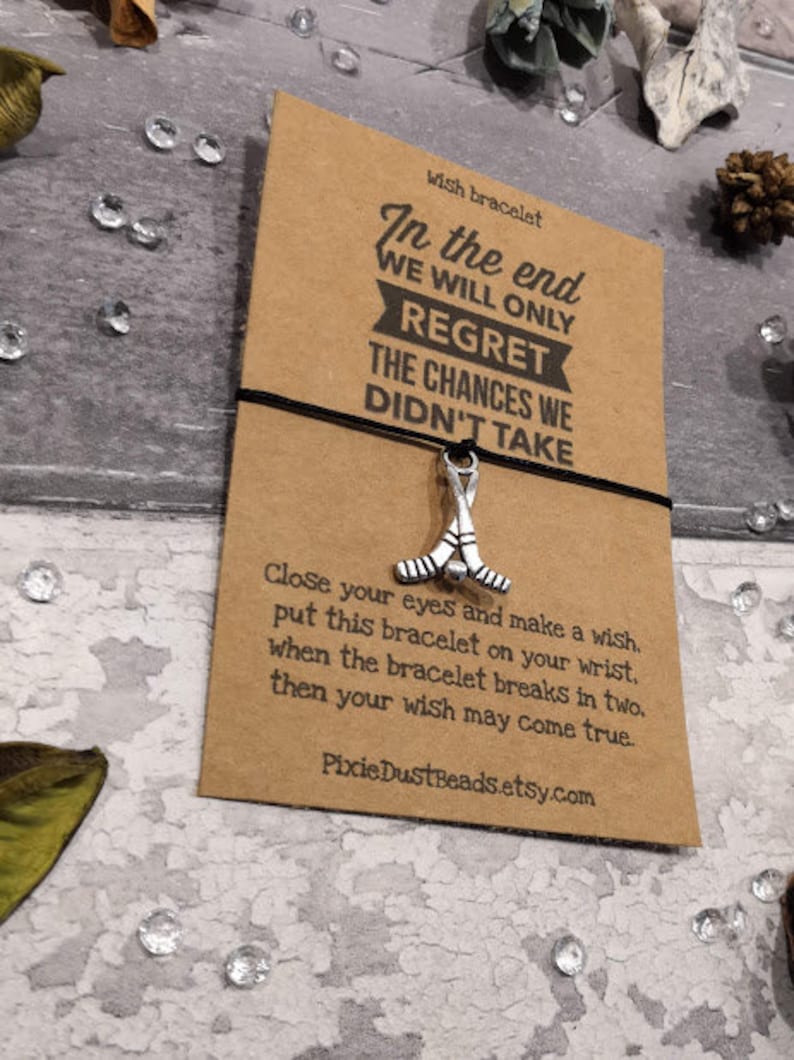 May include: A brown card with the text "In the end we will only regret the chances we didn't take" and a silver hockey stick charm attached to a black string. The card also includes the text "Wish bracelet" and "Close your eyes and make a wish. Put this bracelet on your wrist. When the bracelet breaks in two, then your wish may come true. PixieDustBeads.etsy.com."