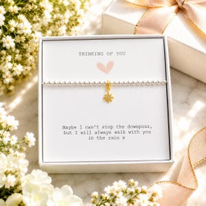 May include: A silver beaded bracelet with a gold star charm featuring a small clear stone, presented in a white gift box. The card inside reads: THINKING OF YOU, a pink heart icon, and: Maybe I can't stop the downpour, but I will always walk with you in the rain x.