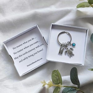 Può includere: Un portachiavi color argento con un ciondolo a forma di tartaruga, un ciondolo con la lettera "M" e una perla blu, presentato in una scatola regalo bianca. La scatola contiene un messaggio stampato. Il portachiavi è su una superficie bianca.