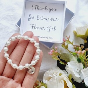 May include: A white pearl bracelet with a silver charm that has the letter "J" on it. The bracelet is in a small white box with the text "Thank you for being our Flower Girl" and the website "artBeadsEtsy.com".
