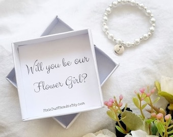 May include: A white box with the text "Will you be our Flower Girl?" and a website address.  A white pearl bracelet with a silver charm is next to the box.  The bracelet has a single letter charm.