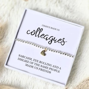 May include: A silver beaded bracelet with a gold heart charm, in a white gift box. The box reads "CHANCE MADE US colleagues" and "SARCASM, EYE ROLLING AND A DISLIKE OF THE SAME PEOPLE MADE US FRIENDS."