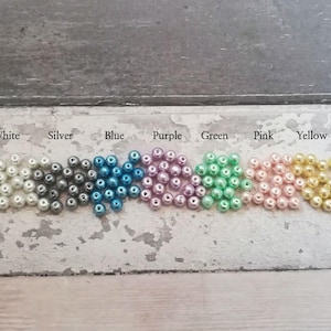 May include: A row of small, round beads in various colors, including white, silver, blue, purple, green, pink, and yellow. The beads are arranged in a line on a light gray surface.