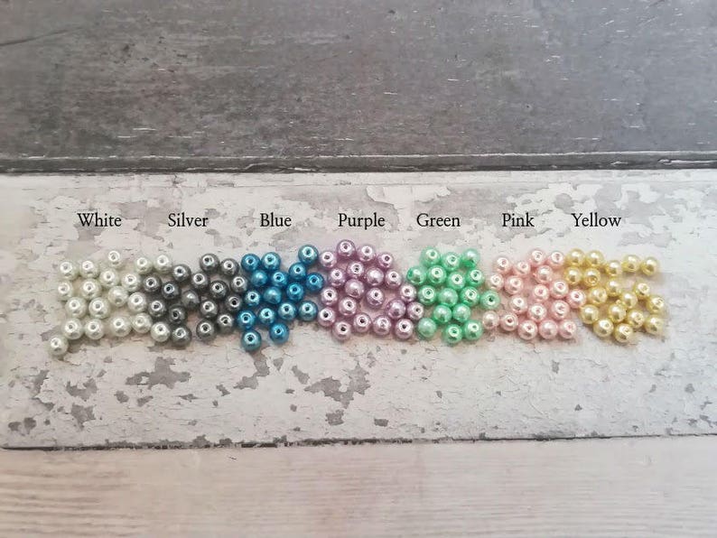 May include: A row of small, round beads in various colors, including white, silver, blue, purple, green, pink, and yellow. The beads are arranged in a line on a light gray surface.