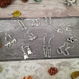 May include: A collection of 12 pairs of silver Halloween-themed earrings. The earrings feature various designs, including bats, ghosts, skeletons, cats, brooms, and teeth.