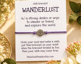 May include: A purple string bracelet with a small silver-toned circular charm featuring a compass design. The bracelet is attached to a white card printed with the text WANDERLUST and instructions for making a wish.
