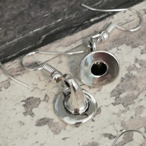 May include: A pair of silver earrings with a hat design. The hats are round and have a small hole in the center. The earrings are hanging from silver hooks.