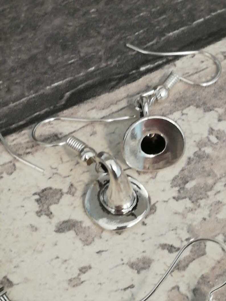 May include: A pair of silver earrings with a hat design. The hats are round and have a small hole in the center. The earrings are hanging from silver hooks.