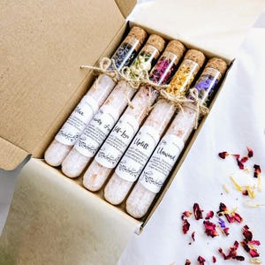 Puede incluir: Una caja de regalo de cart&oacute;n que contiene seis tubos de vidrio con tapones de corcho, llenos de flores secas. Debajo hay seis tubos de sal con etiquetas que dicen "Relax", "Serenity", "Self-Love", "Uplift" y "Unwind". La caja est&aacute; sobre una superficie blanca con p&eacute;talos de flores esparcidos.