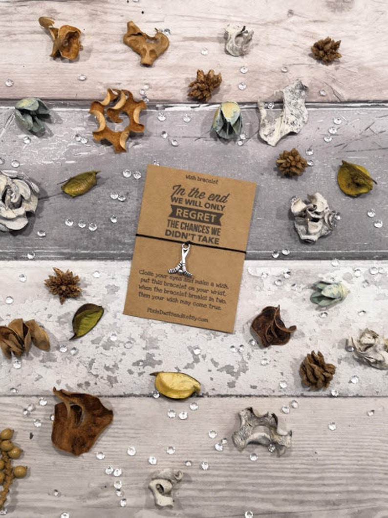 May include: A brown card with the text "In the end we will only regret the chances we didn't take."  The card is attached to a silver charm bracelet with a wishbone charm. The card is surrounded by dried flowers and leaves.