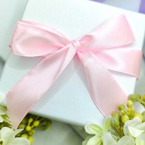 May include: A white gift box with a pink satin ribbon tied in a bow. The box is surrounded by white flowers with green leaves.