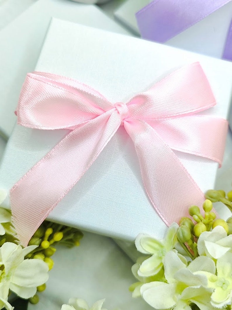 May include: A white gift box with a pink satin ribbon tied in a bow. The box is surrounded by white flowers with green leaves.