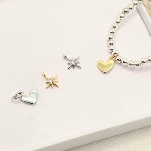 Puede incluir: Una colecci&oacute;n de dijes de joyer&iacute;a y una pulsera de cuentas. Los dijes incluyen un coraz&oacute;n dorado, un coraz&oacute;n plateado y dos dise&ntilde;os de estrella, uno dorado y otro plateado. La pulsera presenta cuentas plateadas y un dije de coraz&oacute;n dorado.