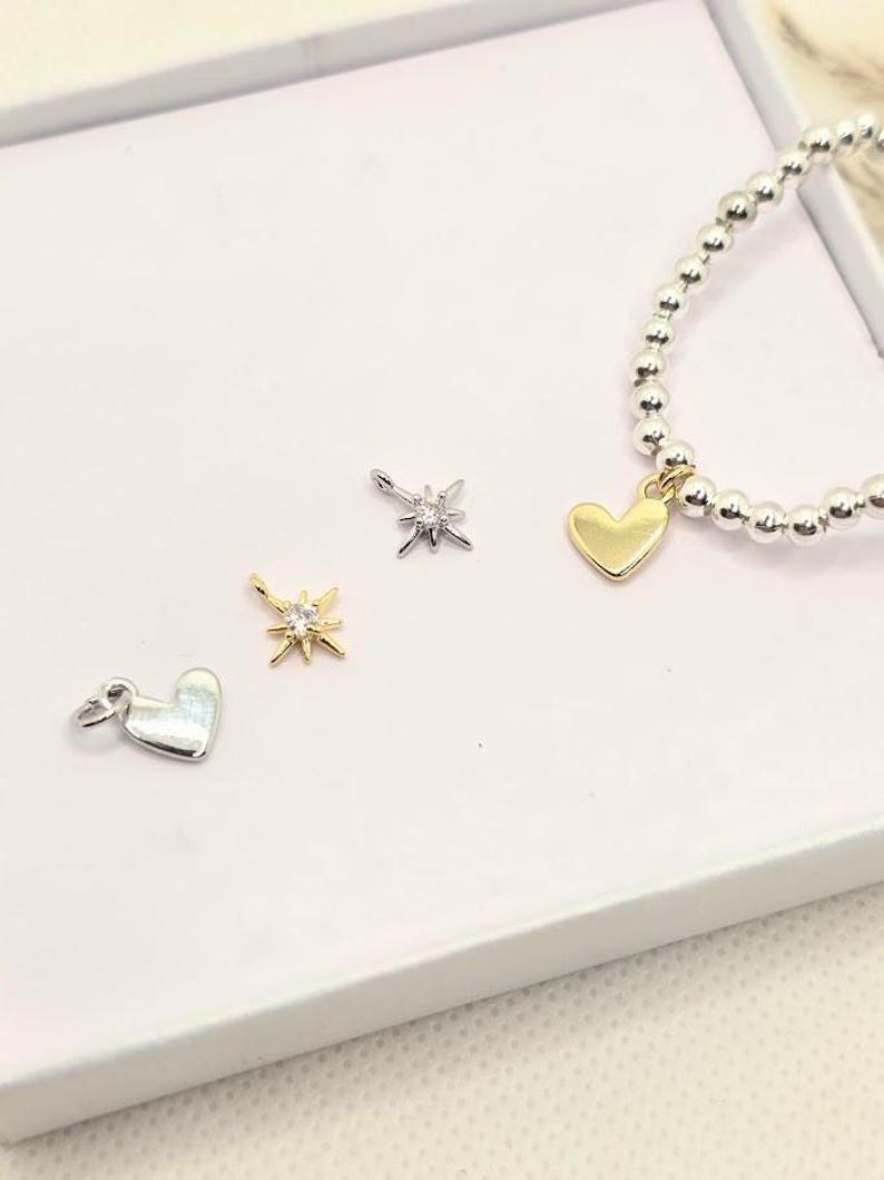 Puede incluir: Una colecci&oacute;n de dijes de joyer&iacute;a y una pulsera de cuentas. Los dijes incluyen un coraz&oacute;n dorado, un coraz&oacute;n plateado y dos dise&ntilde;os de estrella, uno dorado y otro plateado. La pulsera presenta cuentas plateadas y un dije de coraz&oacute;n dorado.
