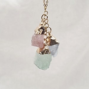 Pode incluir: Um colar de corrente de ouro com três pingentes de cristal de diferentes cores. Os pingentes são rosa, azul claro e verde.