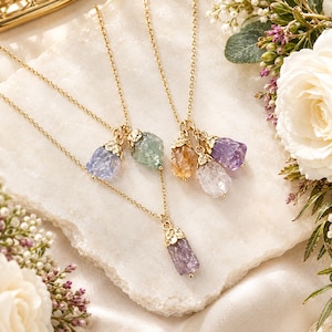 Puede incluir: Dos collares dorados con colgantes de piedras preciosas en bruto sin tallar. Un collar tiene un cristal morado, mientras que el otro presenta un grupo de tres cristales en tonos azul claro, verde, amarillo, transparente y morado, con tapas doradas.