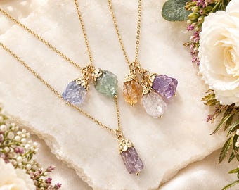May include: Two gold-tone necklaces featuring raw, uncut gemstone pendants. One necklace has a single purple crystal, while the other features a cluster of three crystals in light blue, green, yellow, clear, and purple hues, all with gold-tone caps.