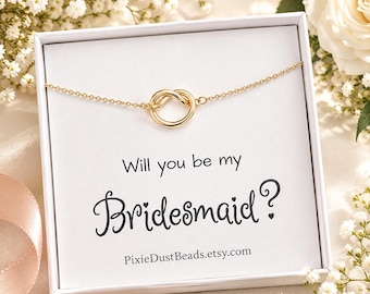May include: A gold-toned necklace with a circular knot pendant centered in a white gift box. The box features the black printed text Will you be my Bridesmaid? and the URL PixieDustBeads.etsy.com on a white card insert.