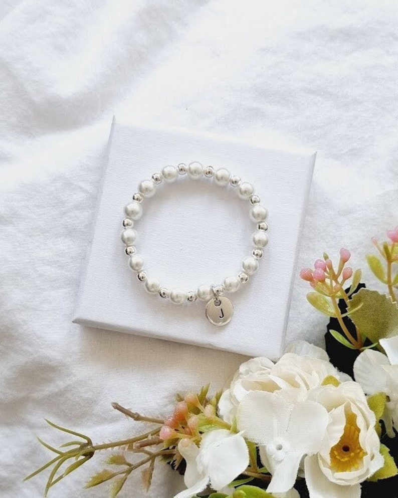 May include: A white pearl bracelet with a silver charm that has the letter "J" on it. The bracelet is on a white surface with flowers in the background.