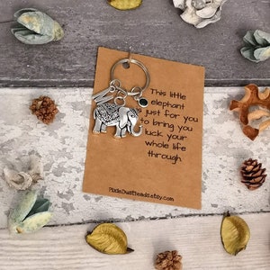 Pu&ograve; includere: Un ciondolo a forma di elefante d'argento attaccato a un portachiavi con una carta marrone su cui &egrave; scritto "This little elephant just for you to bring you luck your whole life through."  PixieDustBeadsEtsy.com &egrave; stampato in fondo alla carta.