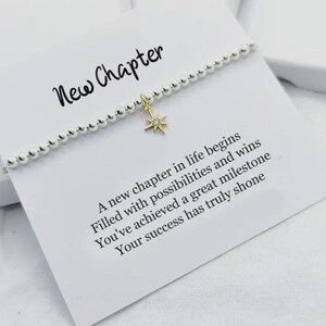 May include: A silver beaded bracelet with a gold starburst charm rests on a white card. The card features the words "New Chapter" in cursive, along with an inspirational message. This is a delicate piece of jewellery.