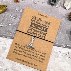 May include: A brown card with the text "In the end we will only regret the chances we didn't take."  The card is tied with a black string with a silver hockey stick charm. The card also includes instructions for making a wish with the bracelet.