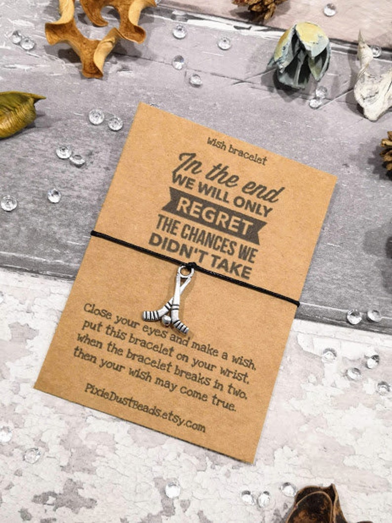 May include: A brown card with the text "In the end we will only regret the chances we didn't take."  The card is tied with a black string with a silver hockey stick charm. The card also includes instructions for making a wish with the bracelet.