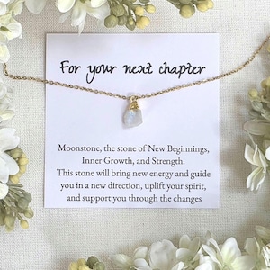 Puede incluir: Un delicado collar dorado con un colgante de piedra lunar, exhibido en una tarjeta con el texto "For your next chapter". La tarjeta también incluye texto sobre las propiedades de la piedra lunar. Flores blancas rodean la tarjeta.