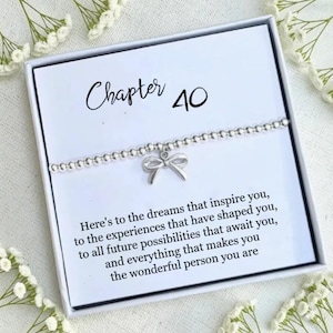 May include: A silver beaded bracelet with a bow charm, presented in a white box. The box has the text "Chapter 40" and an inspirational message. White flowers surround the box.